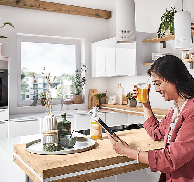 Une femme savoure son thé du matin tout en naviguant sur une tablette dans une cuisine moderne et lumineuse avec des accents en bois élégants et des plantes vertes.
