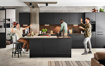 Tre uomini si godono un incontro informale e divertente in una cucina moderna con mobili neri eleganti, preparando cibo e condividendo risate.