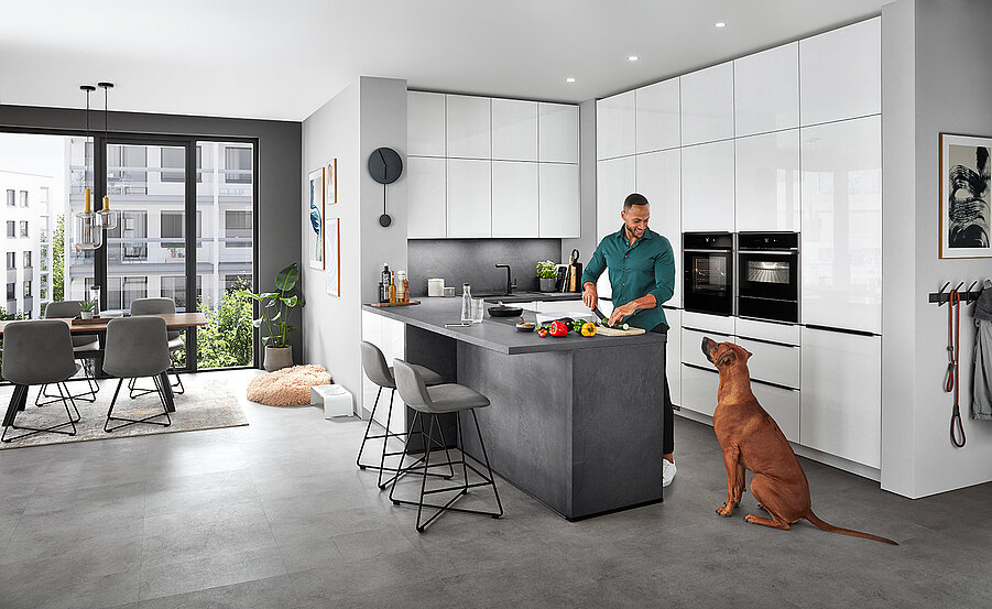Een modern keukeninterieur met een man die eten bereidt op het aanrecht terwijl een hond in de buurt zit, met strakke apparaten en een goed verlichte eethoek.