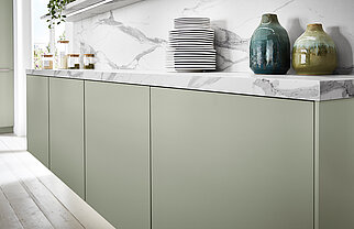 Cucina moderna con linee pulite caratterizzata da armadietti verde salvia, piani in marmo e un'organizzazione minimalista di piatti e vasi in ceramica.