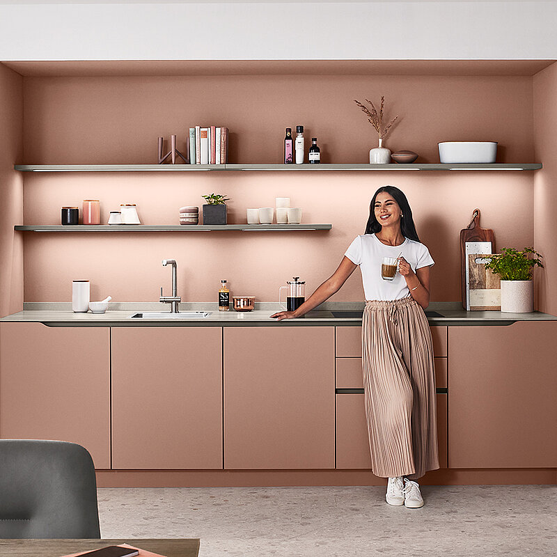 Conception de cuisine moderne avec une personne debout près du comptoir, mettant en vedette des armoires élégantes de couleur rose poudré, des appareils élégants et des étagères ouvertes avec une décoration minimaliste.