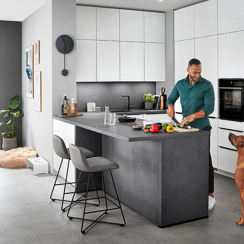 Een stijlvolle moderne keukenscène met een man die eten bereidt op een kookeiland terwijl een hond toekijkt, wat een comfortabele, eigentijdse huiselijke levensstijl weerspiegelt.