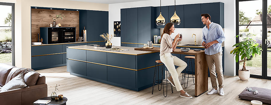 Een moderne keuken met elegante blauwe kasten en gouden armaturen, waar twee personen genieten van een warm gesprek onder het genot van drankjes in een stijlvolle, huiselijke ruimte.
