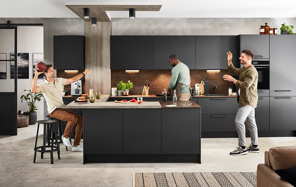 Tres hombres disfrutan de una reunión casual y divertida en una cocina moderna con elegantes gabinetes negros, preparando comida y compartiendo risas.