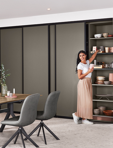 Een eigentijdse eetkamer met een glimlachende vrouw naast een open wandrek, met stijlvol meubilair en een minimalistisch kleurenschema.