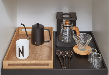Una estación de café elegantemente dispuesta con una tetera negra, molinillo, cristalería y cucharas únicas en una bandeja de madera elegante, mostrando un diseño moderno de utensilios de cocina.