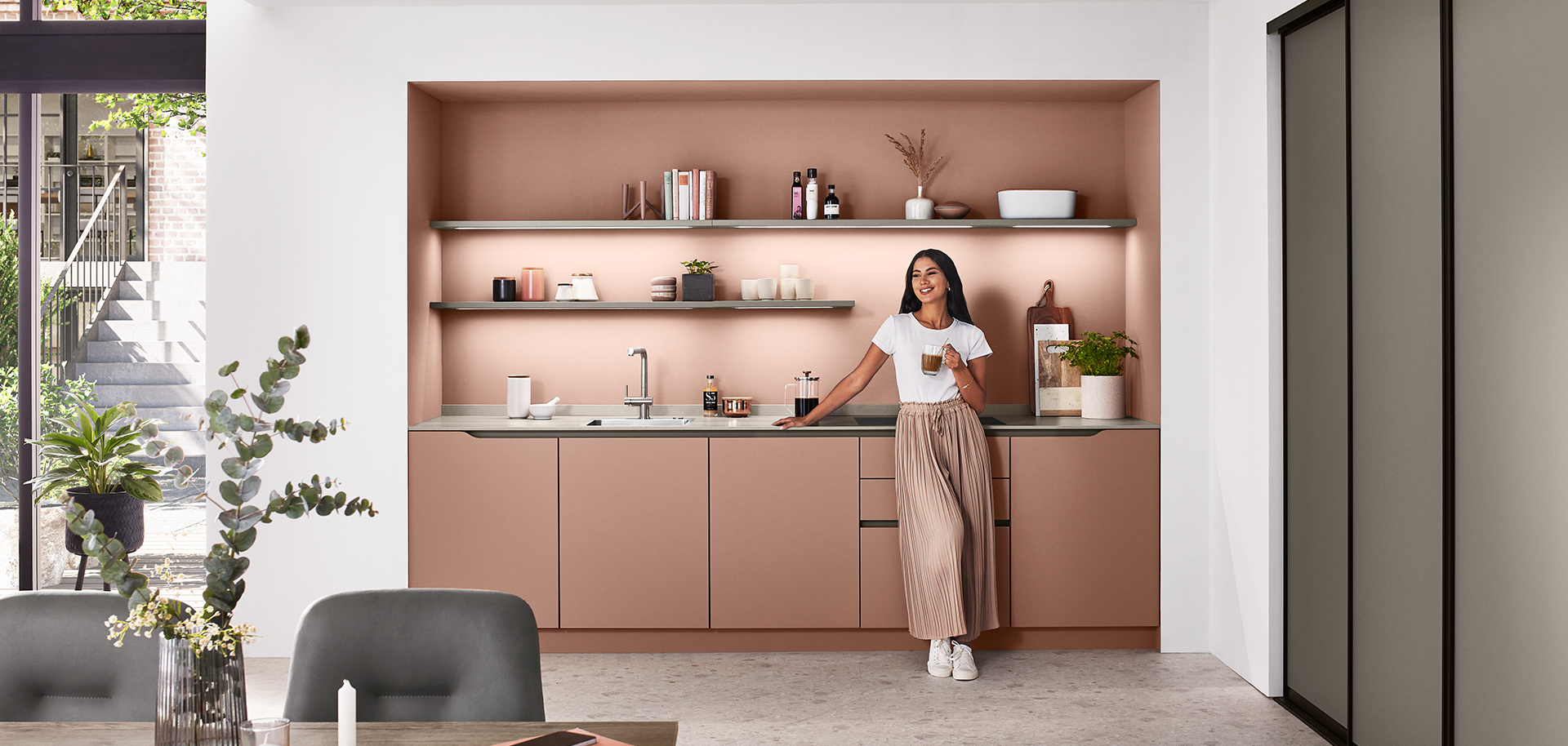 Conception de cuisine moderne avec une personne debout près du comptoir, mettant en vedette des armoires élégantes de couleur rose poudré, des appareils élégants et des étagères ouvertes avec une décoration minimaliste.