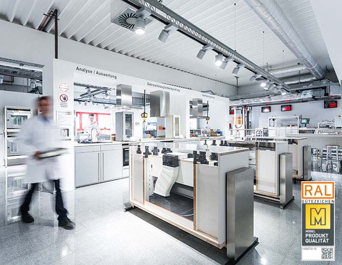A modern, clean laboratory with stainless steel workstations, technical equipment, and a blurred figure of a professional in a lab coat in motion.