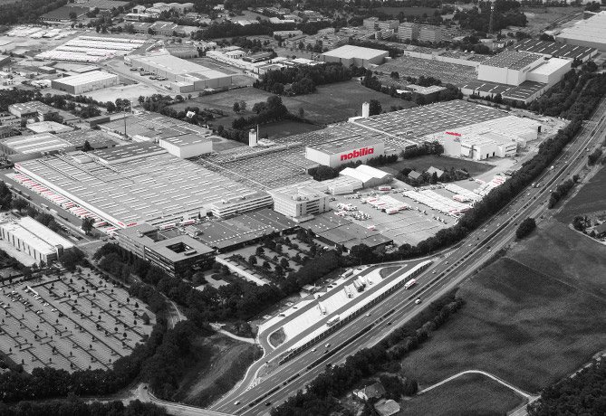 Aerial grayscale photo of an industrial complex with highlighted "Mobilia" brand, showcasing a large warehouse, parking lots, and access roads amid green surroundings.