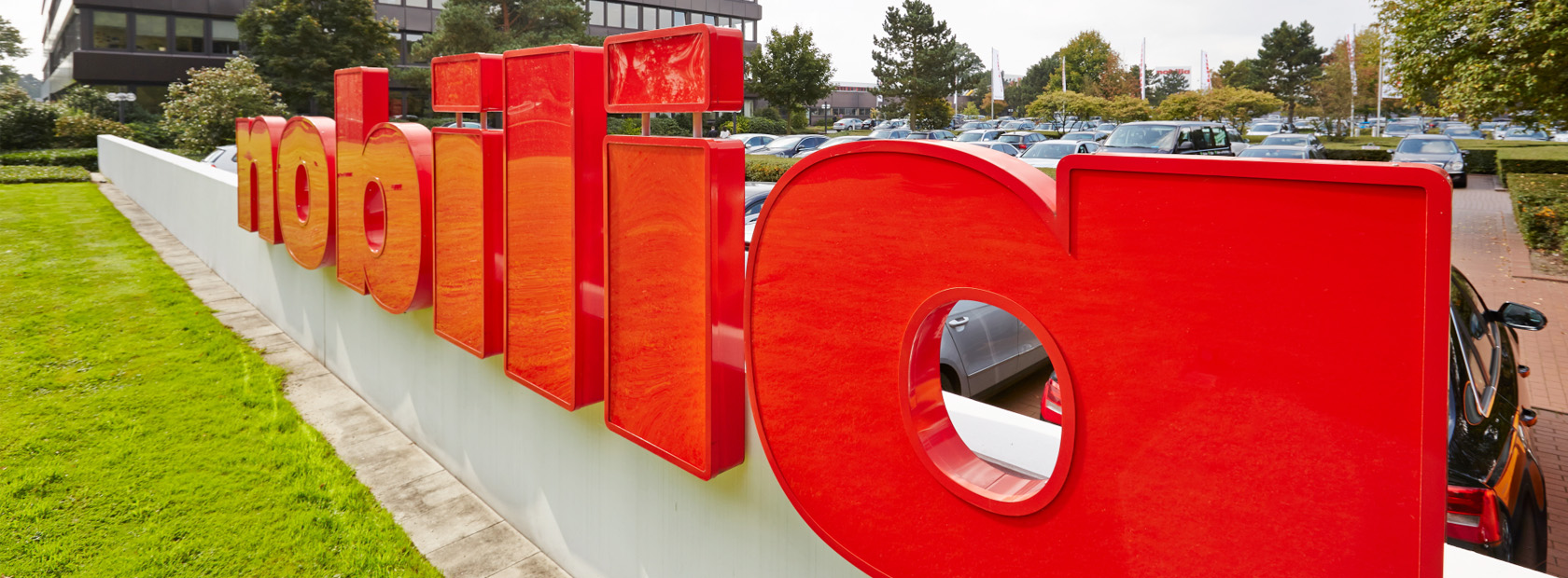 Rote dreidimensionale Buchstabenschilder, die ein Wort bilden, im Freien installiert, mit üppigem Grün und einem modernen Gebäude im Hintergrund.