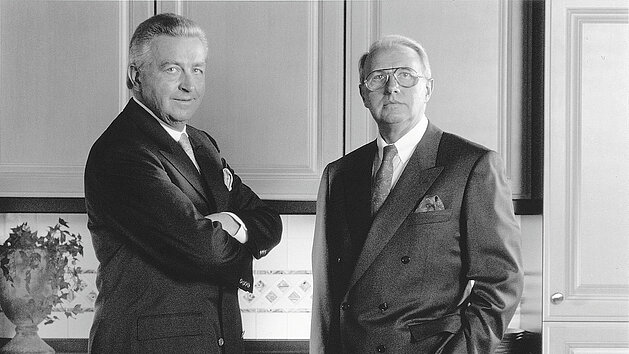 Zwei elegante Männer in Anzügen posieren vor stilvollen Küchenschränken im Jahr 1965. nobilia im Hintergrund sichtbar.