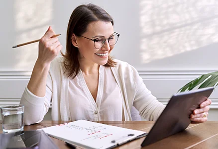 Lächelnde Frau mit Brille und Stift in der Hand, schaut auf ein Tablet, neben ihr liegt ein Notizblock.