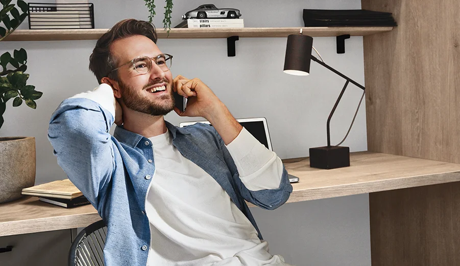 Eiche Montreal Nachbildung (126) Ein zufriedener junger Berufstätiger unterhält sich auf seinem Smartphone, während er bequem in einem stilvollen Homeoffice mit minimalistischer Einrichtung und stimmungsvoller Beleuchtung sitzt.