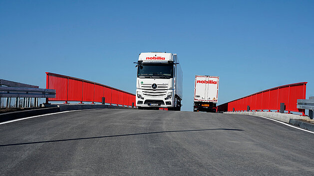 weißer Lastwagen von nobilia fährt auf einer roten Brücke -- hinter ihm steht ein weiterer Lastwagen -- blauer Himmel im Hintergrund